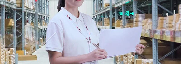 Employee in a white uniform holding paperwork while standing in a warehouse aisle filled with shelves and boxes.