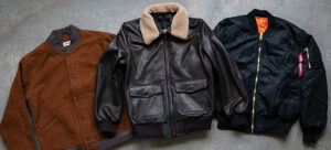 Three men’s bomber jackets laid flat on a concrete surface, including a brown wool blend, a black leather with shearling collar, and a black nylon style with zipper and orange lining