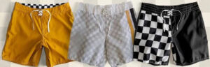 Three pairs of women's boardshorts in yellow, light gray, black and white checkered pattern, and solid black, laid flat with lace-up waistbands.