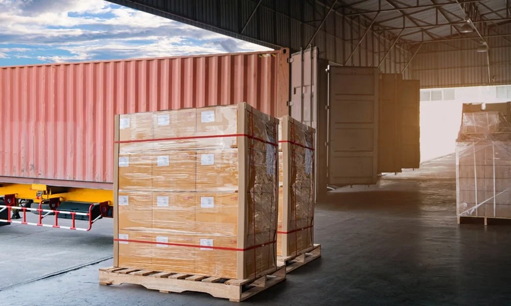 Pallets of packaged goods being loaded into a shipping container inside warehouse.