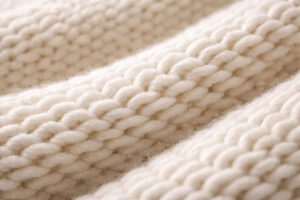 Close-up of soft cream cotton knit fabric featuring chunky braided weave texture, showcasing warmth, comfort, and premium craftsmanship for sweaters and winter apparel.