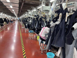 Large-scale denim manufacturing factory with sewing stations, hanging garment panels, and workers assembling jeans in an industrial apparel production line.
