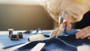 Tailor cutting dark fabric with scissors beside measuring tape and sewing tools during garment manufacturing and apparel production process