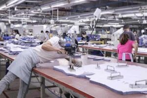 Worker using fabric cutting machine in busy clothing factory with multiple workers in the background.