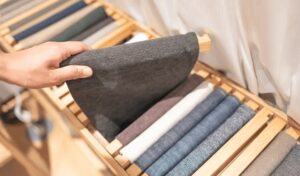 Hand selecting dark fabric roll from organized textile samples displayed on wooden racks in fabric showroom or workshop