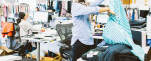 Fashion designers working in a busy studio, reviewing fabrics and clothing samples, with computers and garment racks in the background, illustrating raw material sourcing and apparel development.