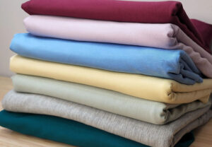 Stack of soft cotton fabrics in pastel and neutral colors neatly folded on a wooden surface, perfect for apparel and textile projects.