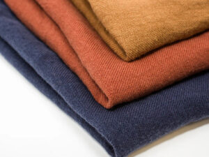 Stack of folded knit fabric swatches in blue, rust, and mustard colors, showcasing soft texture, durability, and premium apparel material quality.