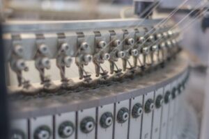 Close-up of an automated circular knitting machine producing fabric with multiple needles and yarn feeds in a modern textile manufacturing facility.