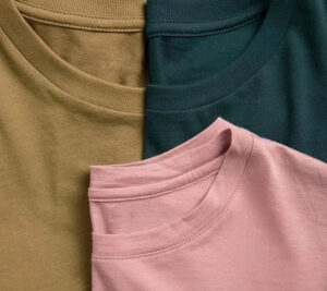 Close-up of three folded Pima cotton T-shirts in olive green, dark teal, and muted pink, highlighting soft fabric texture and neatly stitched crew necklines.