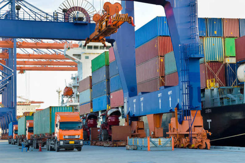 Cargo trucks moving containers in busy port terminal with cranes loading and unloading shipping containers beside docked vessels.
