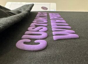 lose-up of a black hoodie with 3D purple puff print lettering on the chest, showing texture.