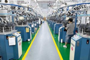 Modern seamless knitting machines aligned inside textile factory with worker operating automated equipment for garment manufacturing production.