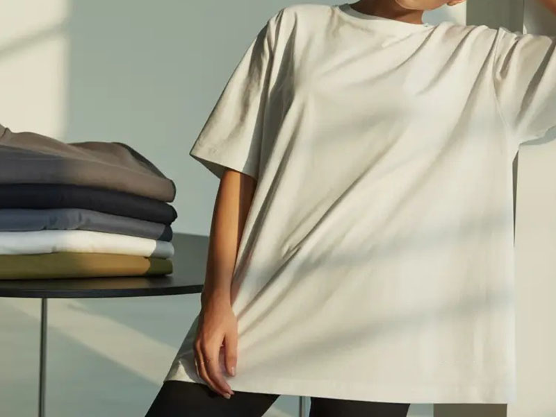Person wearing an oversized plain T-shirt with folded T-shirts in neutral colors stacked on a table nearby.