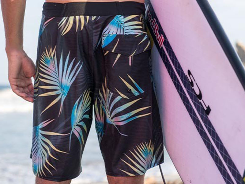 Person wearing tropical leaf-print boardshorts beside a surfboard, showcasing lightweight quick-dry fabric, cargo pocket design, and stylish beachwear for surfing and water sports.