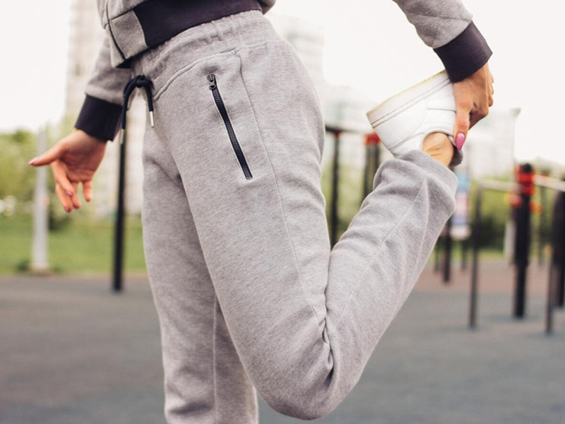Woman stretching leg outdoors wearing gray athletic sweatpants and sneakers.