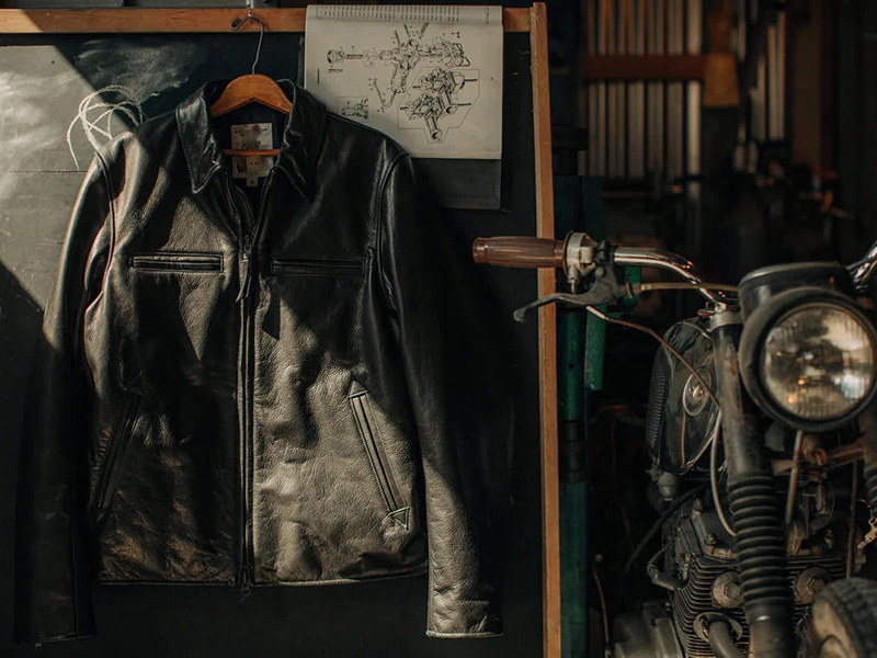 Black leather jacket hanging in a workshop beside a motorcycle, showcasing classic craftsmanship, rugged style, and premium biker-inspired design.