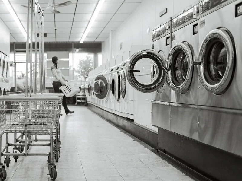 Row of industrial washing machines inside laundromat with customer carrying laundry basket.