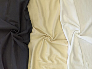 Swatches of cotton fabric in black, beige, and white, twisted to show texture, softness, and natural fibers for T-shirt or garment making.